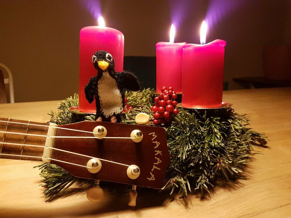 Ukulele vor Adventskranz. Auf der Ukulele sitzt ein Stoffpinguin. Auf dem Adventskranz leuchten Kerzen.