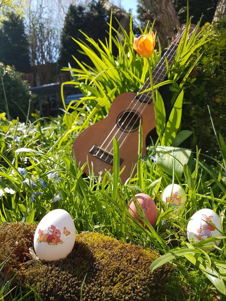Ukulele im Gras vor einem Busch lehnend, daneben eine rote Blume, davor kleine blaue Blumen und bunte Ostereier mit nostaltigischen Osterhasenbildern
