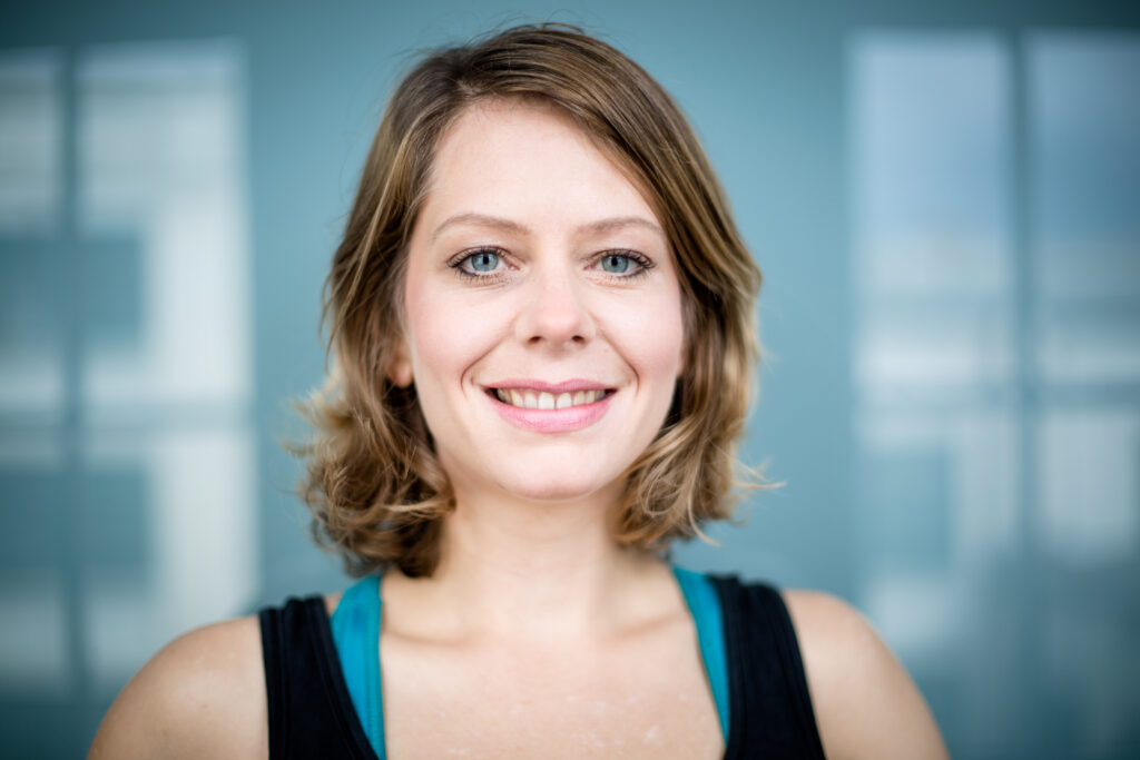 Foto von Svenja Olbrich vor hellblauem Hintergrund. Sie trägt ein blau-schwarzes Top und lächelt in die Kamera.