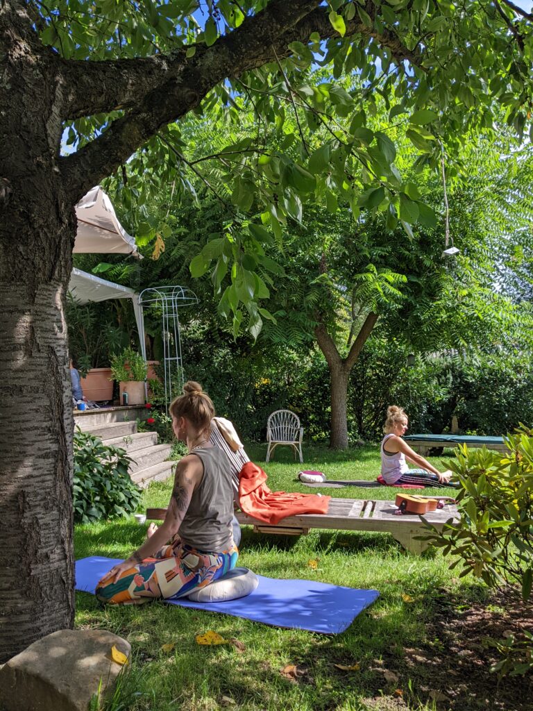 Foto von zwei Teilnehmerinnen des Yogakurses. Sie sitzen auf Matten im Garten. Die Sonne scheint. Drumherum dicke grüne Bäume.
