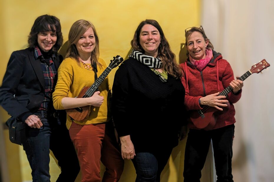 Foto von Joyce Mordoh, Ukulili, Sabina Saracevic und Ariane Schlesinger. Sie stehen vor einer gelben Wand, lächeln in die Kamera und zwei tragen eine Ukulele.