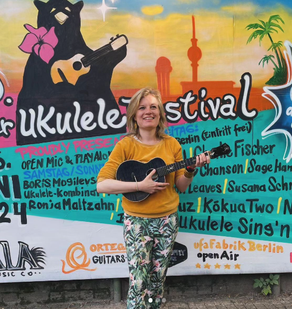 Foto von Ukulili vor Poster des Berliner Ukulele Festivals. Sie trägt offene blonde Haare, einen gelben Pullover, eine bunte Leggings und eine dunkelblaue Ukulele und lacht in die Kamera. Im Hintergrund ein Bild von Berliner Bär und Übersicht von Künstler*innen.