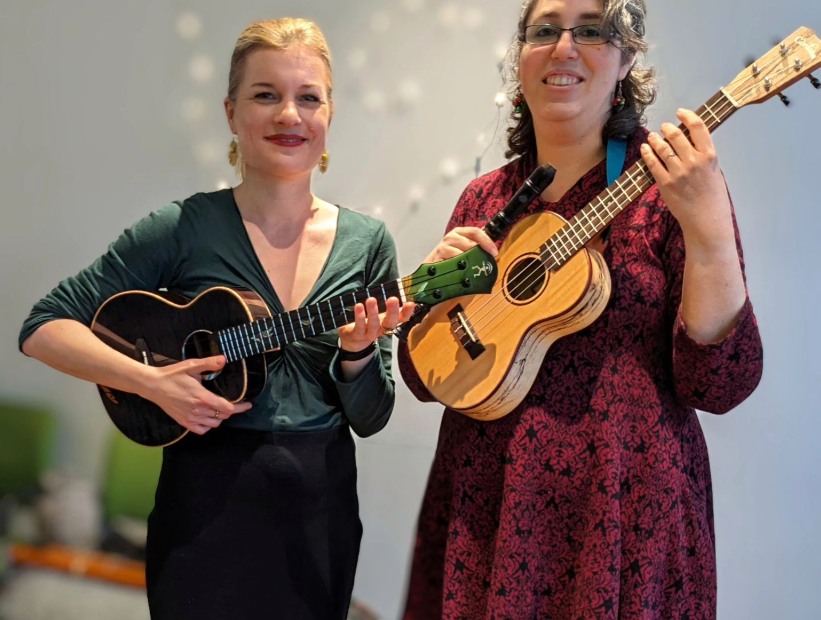 Foto von Ukulili und Maria Gilman. Ukulili trägt schwarzen langen Rock, grünes Oberteil und Ukulele. Maria trägt rotes Kleid und Ukulele uns Flöte.