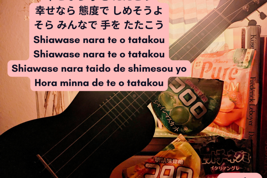 Foto von Ukulele mit japanischen Gummibären in schummrigen Licht. Im Hintergrund bunte Papierlampe und getrocknete Blume. Text: Lied mit japanischen Schriftzeichen. Darunter Shiawase nara te o tatakou. Shiawase nara te o tatakou. Shiawase nara taido de shimesou yo. Hora minna de te o tatakou.